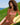 A woman with curly brown hair relaxes on a yellow lounge chair on a grassy hill overlooking the blue ocean. She is wearing a red and white gingham bikini.